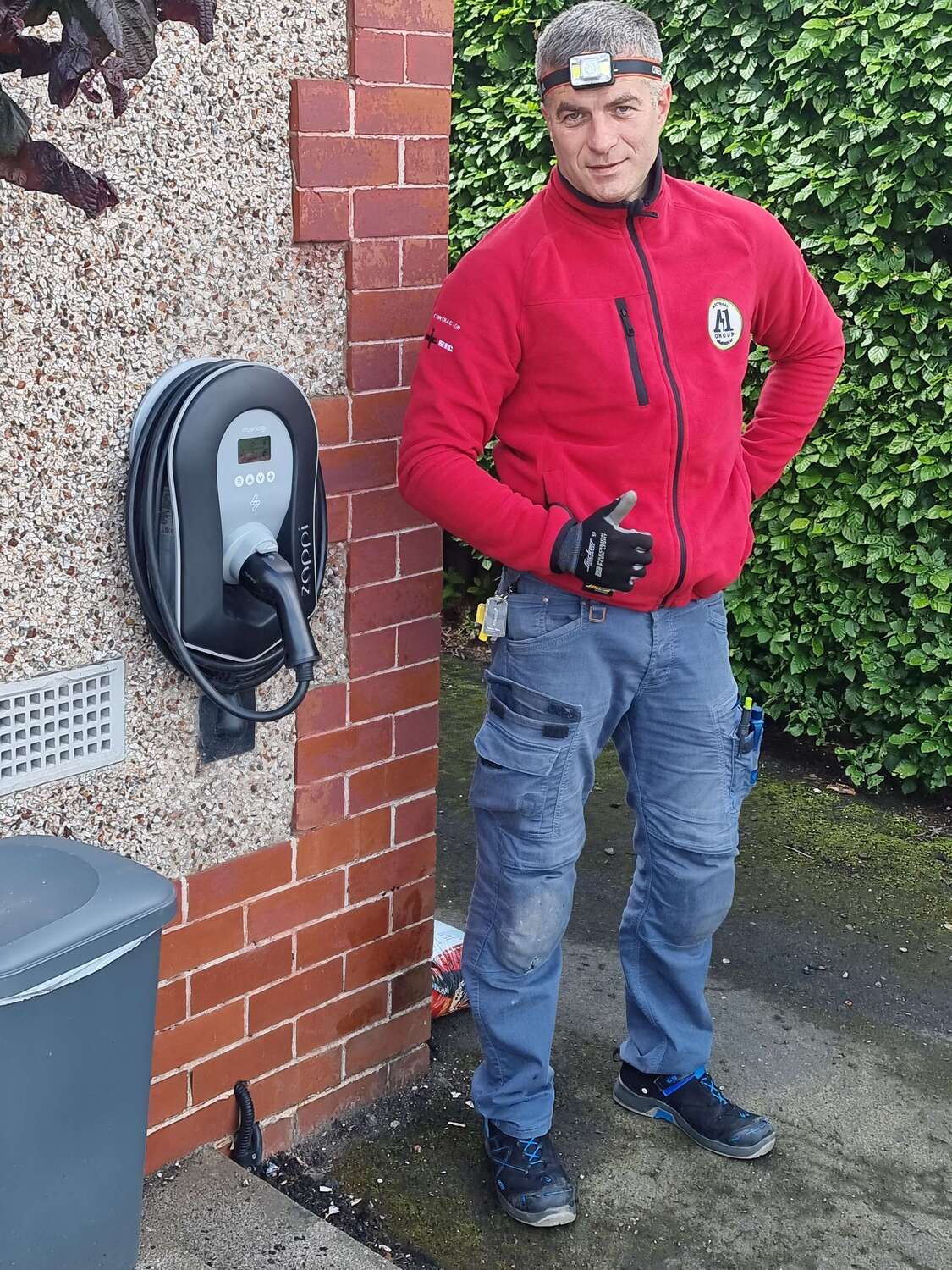 Electrician in red jacket giving a thumbs up next to a newly installed EV charger on an exterior brick wall.