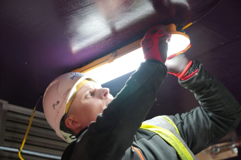 An electrician wearing a white hard hat, safety vest, and red gloves is installing or adjusting a bright overhead light fixture in an industrial setting.