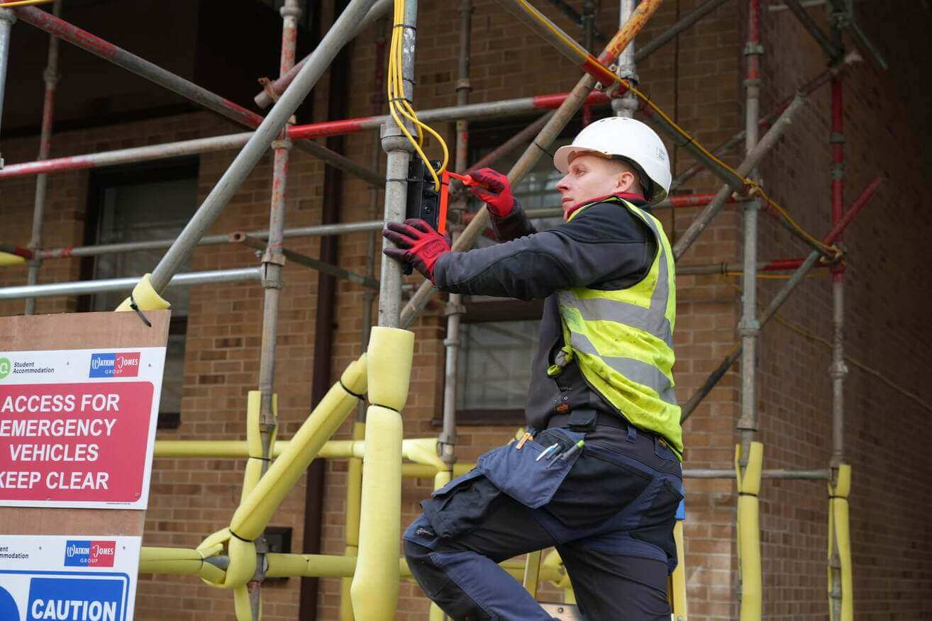 Electrician in a hard hat and safety vest working on wiring on a scaffold