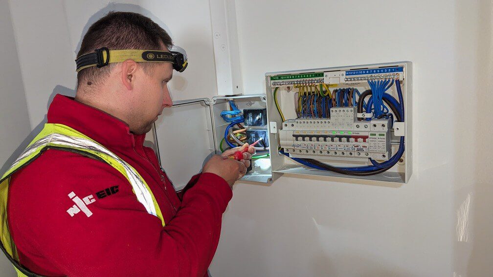 NICEIC certified electrician wearing a headlamp and safety vest working on an electrical panel