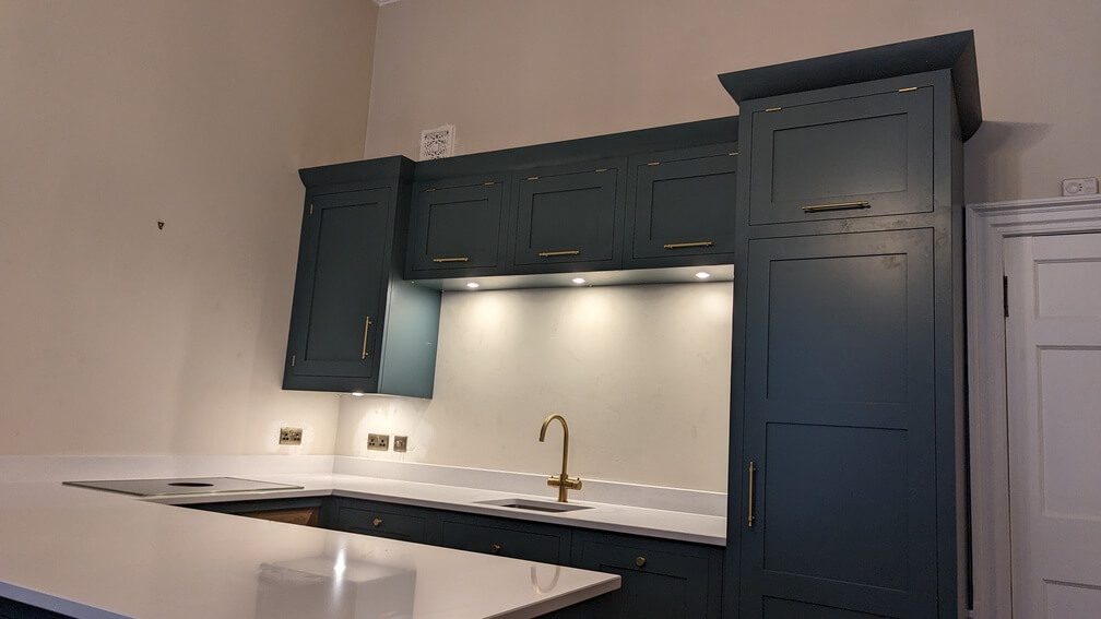 A modern kitchen with dark green cabinets, brass hardware, and white countertops. The kitchen features a gold faucet, built-in appliances, and under-cabinet lighting.