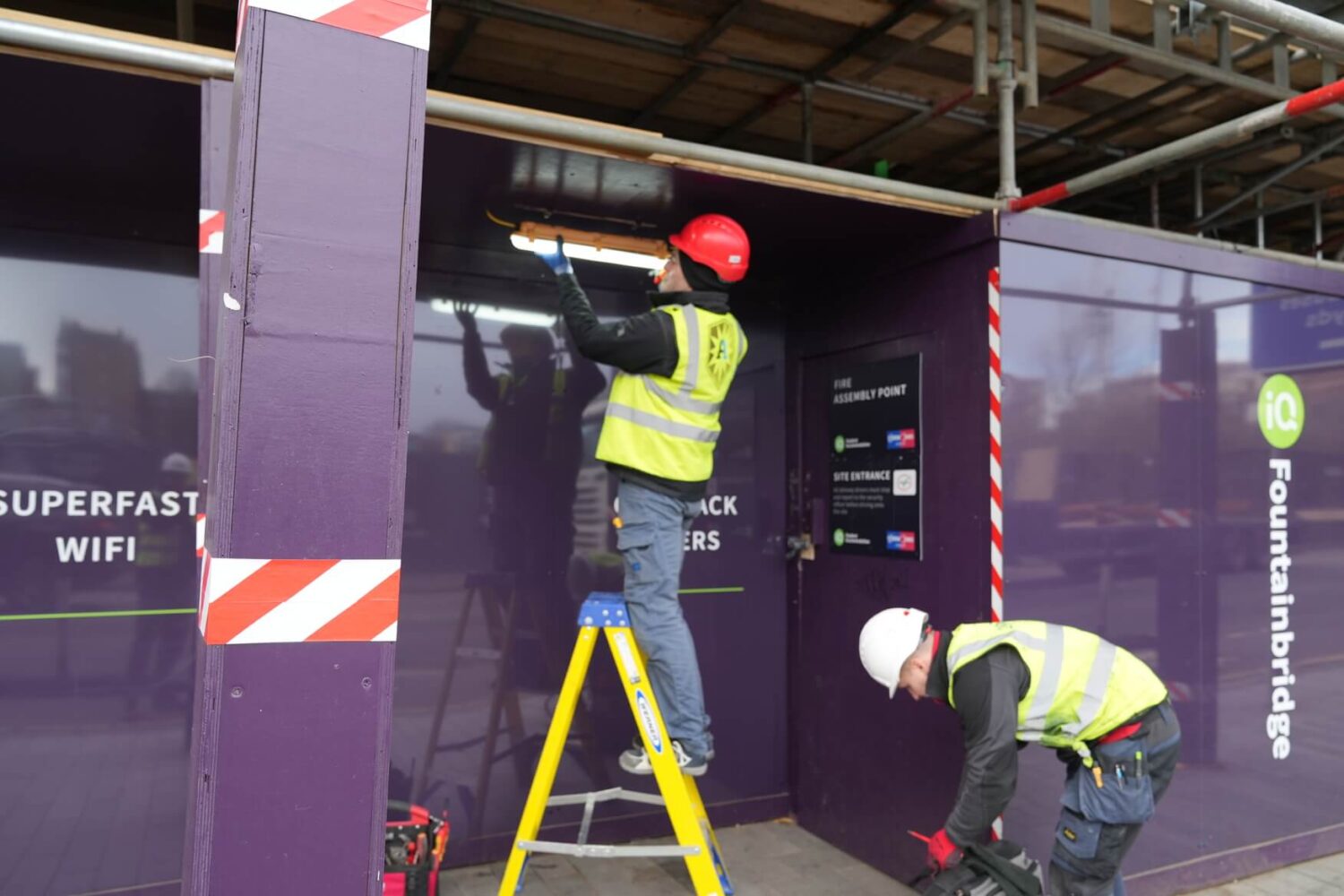 Two commercial electricians in safety gear working on a construction site, one on a ladder installing a light fixture